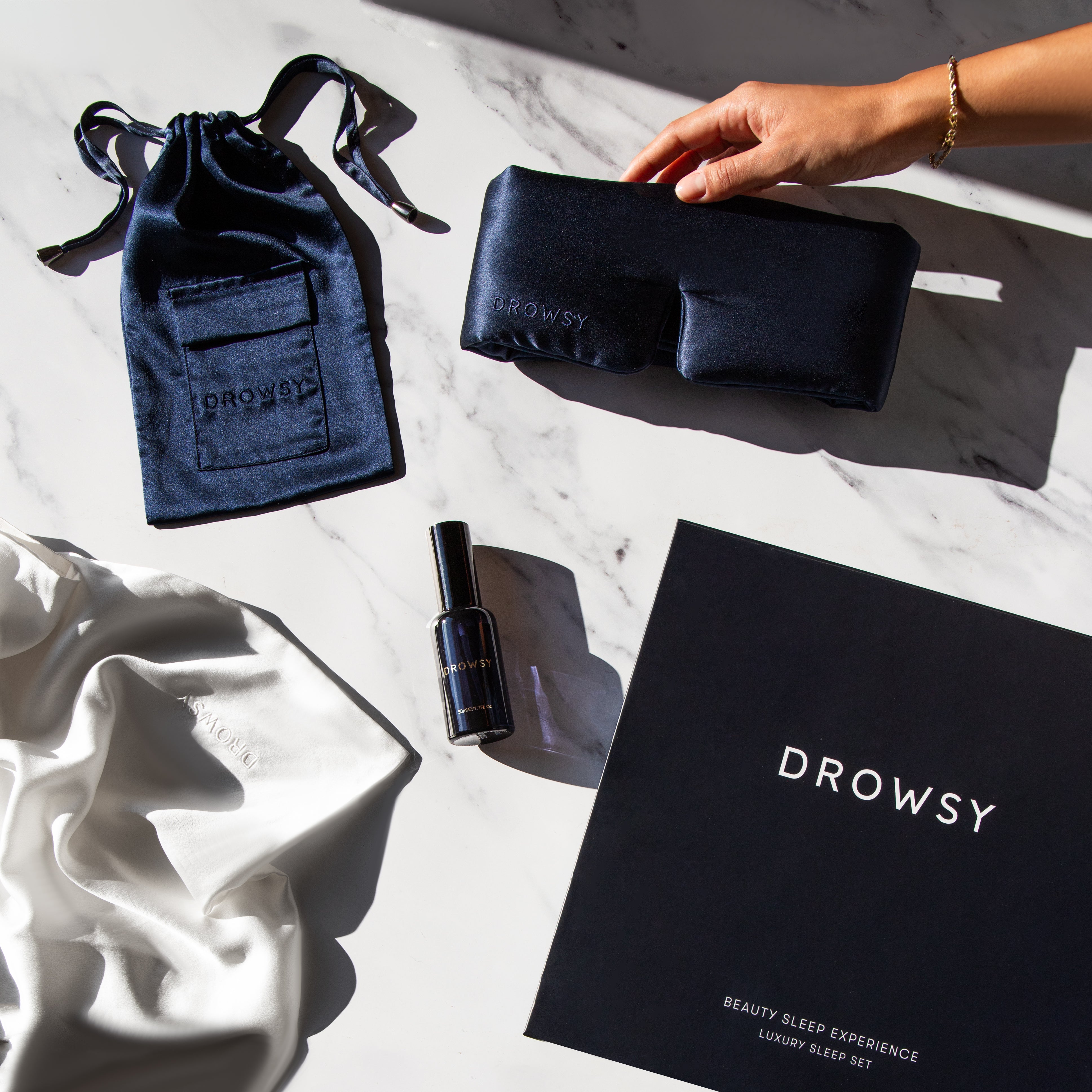 A hand reaches for a navy blue Drowsy sleep mask on a marble surface. Nearby are a matching drawstring pouch, a satin pillowcase, a spray bottle, and a black Drowsy gift box.