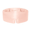 A woman with curly hair stands against a white background, wearing the us.drowsysleepco Sunset Pink silk eye mask and matching pajamas—ready for beauty sleep.