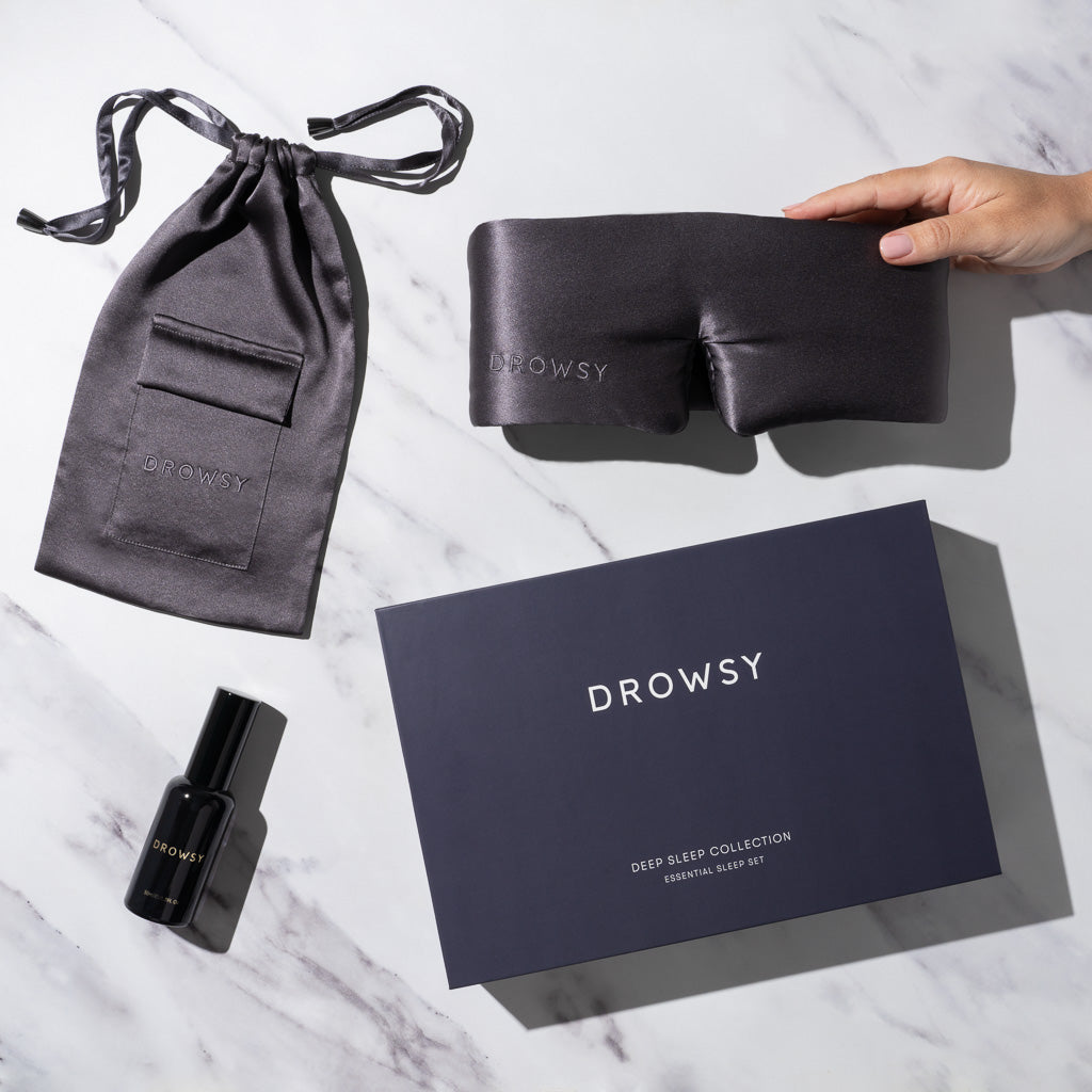 A hand holds a luxury silk sleep mask with a matching pouch, pillow spray, and a dark blue us.drowsysleepco box labeled 