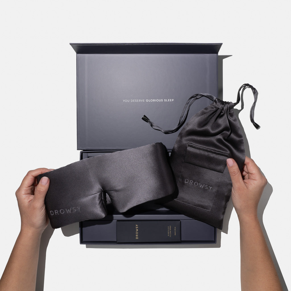 Two hands hold the Moonlight Shadow - Deep Sleep Collection by us.drowsysleepco: a luxury silk sleep mask and pouch above a gift box with deep sleep pillow spray. Inside the lid reads 