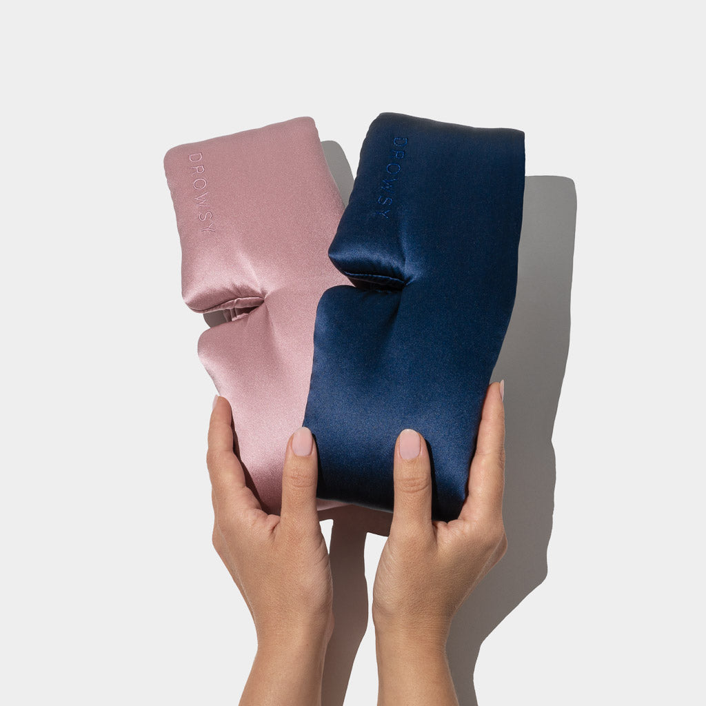 Two hands hold the Drowsy Lovers Pack, featuring a pink and a navy blue luxury silk sleep mask embossed with 