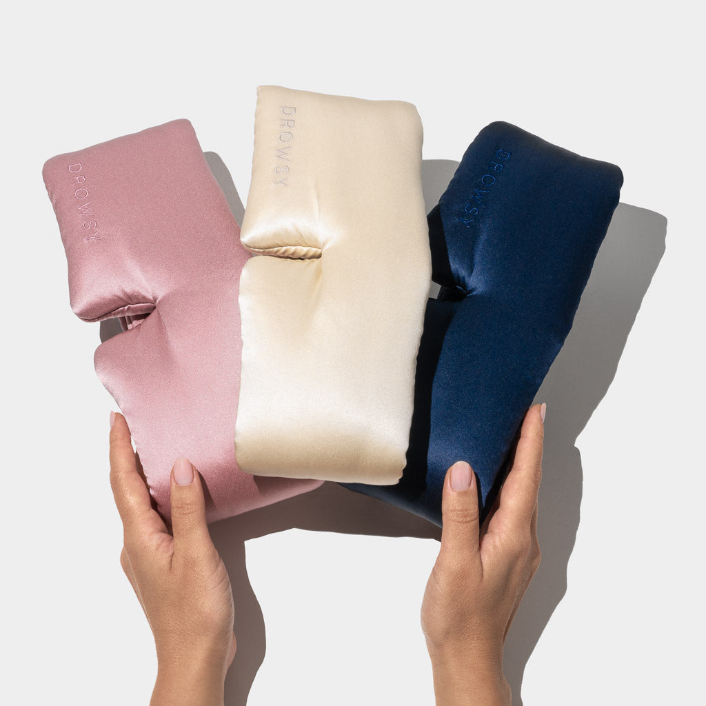Two hands hold three Drowsy Besties Pack luxury masks in pink, cream, and navy blue. Crafted from mulberry silk and embossed with the brand name, they sit side by side on a light background—perfect for beauty sleep.