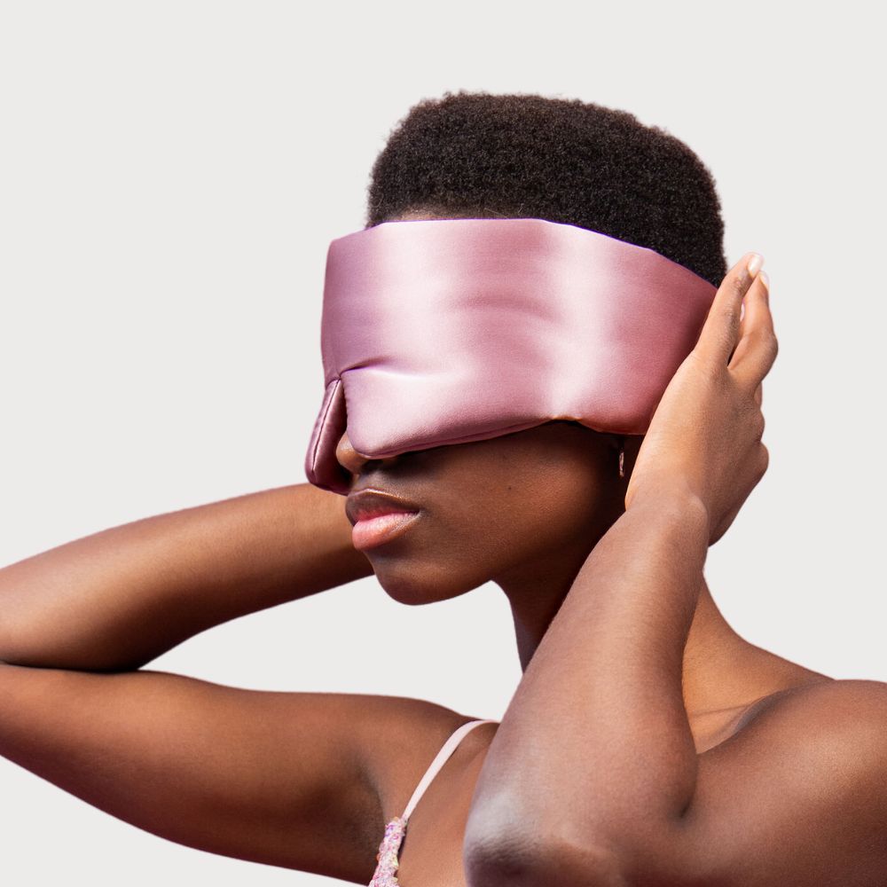 A person with short hair wears a shiny, light pink satin eye mask, covering their eyes and part of their nose. They hold the mask in place with both hands against a plain, light background.