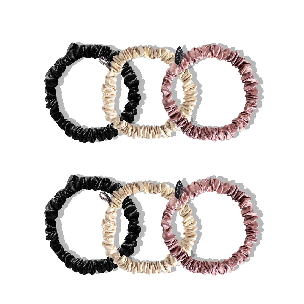 Six scrunchies made of shiny fabric in black, beige, and pink colors, arranged in two rows on a white background. Each row contains one scrunchie of each color.