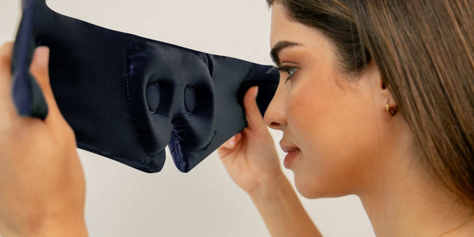 A woman holds a navy blue sleep mask with contoured eye cups, preparing to put it on. She is shown from the side with the mask close to her face.
