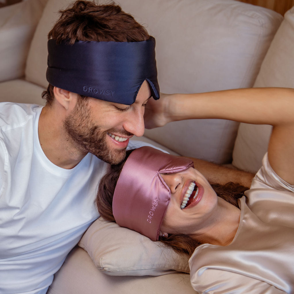 A smiling man and woman relax on a couch, both wearing silk eye masks. The man wears a navy eye mask and a white shirt, while the woman wears a pink eye mask and a satin top, laughing as she leans back comfortably.