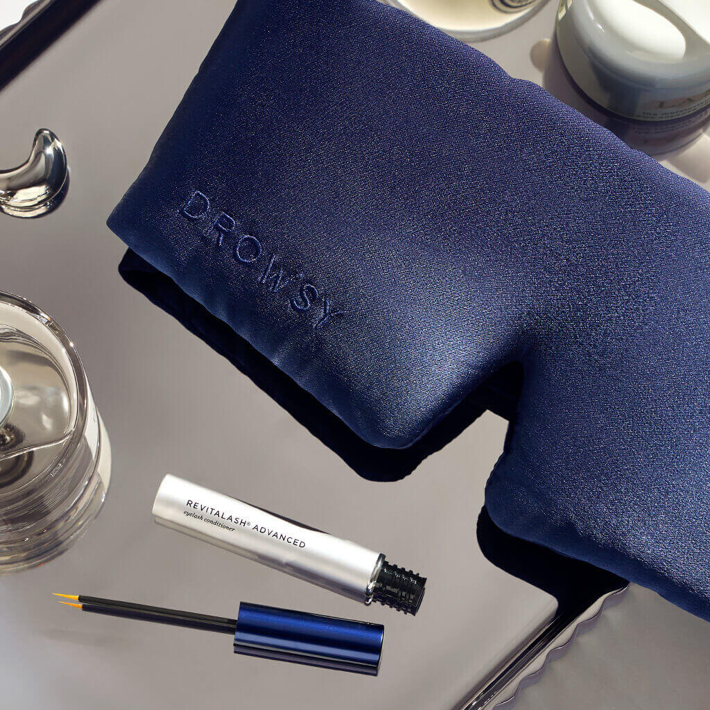 A Drowsy navy sleep mask and a RevitaLash Advanced eyelash conditioner from the RevitaLash® Cosmetics x Drowsy® Dream Lashes Kit rest on a reflective surface beside a glass of water and a cream jar.