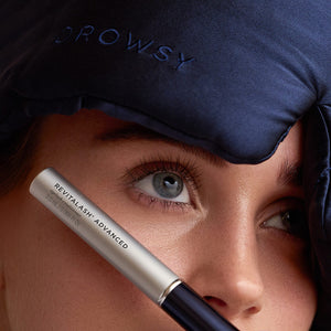 A close-up of a person wearing a navy blue Drowsy sleep mask and holding the RevitaLash® Cosmetics x Drowsy® Dream Lashes Kit near their eye, highlighting long lashes.