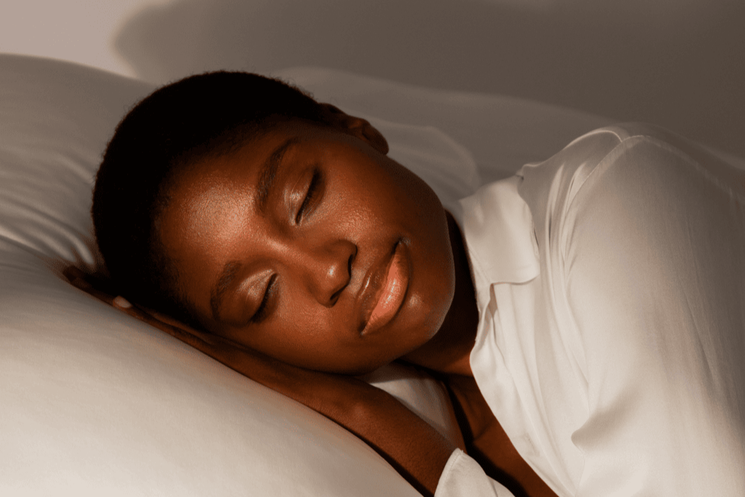 A woman with short hair and closed eyes peacefully rests her head on a white pillow, wearing a white shirt, and bathed in soft, warm light.