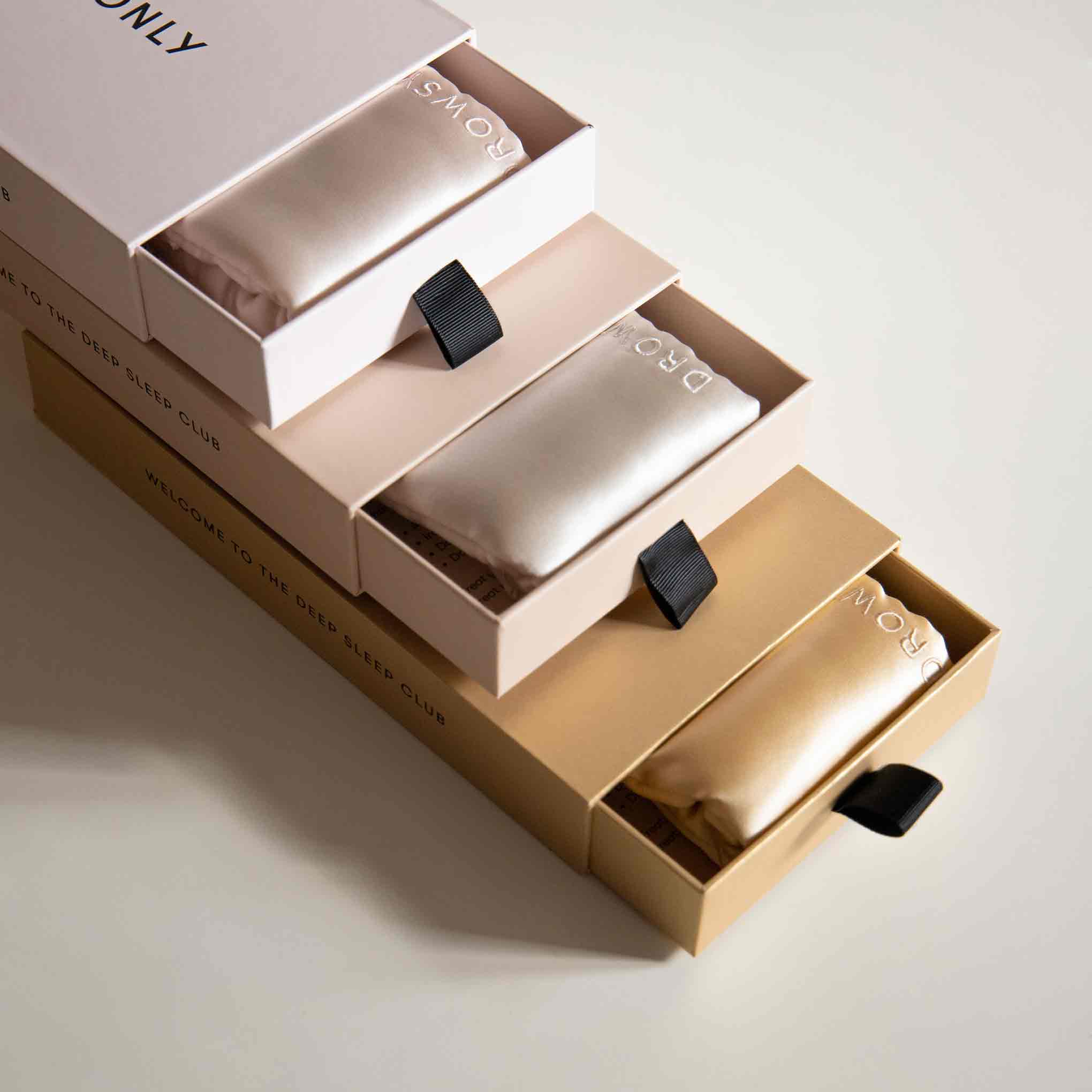 Three rectangular boxes in soft beige and white tones are stacked on a light surface, each partially open to reveal silky fabric pouches with black pull tabs and the word “DROWSY” printed on top.