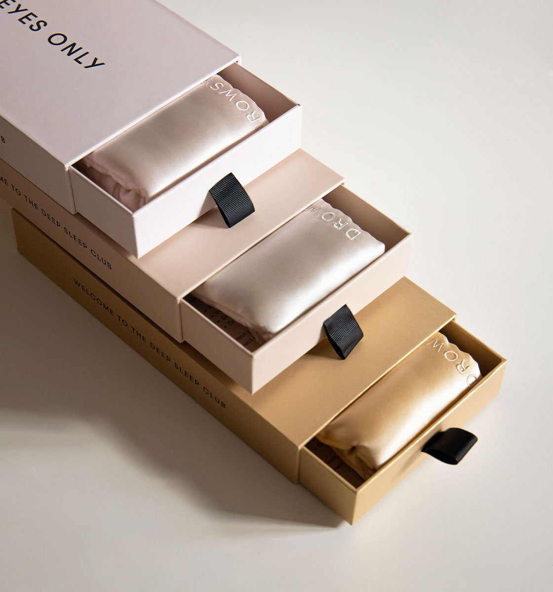 Three satin sleep masks in rose, cream, and gold, each with a black ribbon loop, are presented in matching colored boxes partially opened to reveal the masks. The boxes are stacked diagonally on a neutral background.