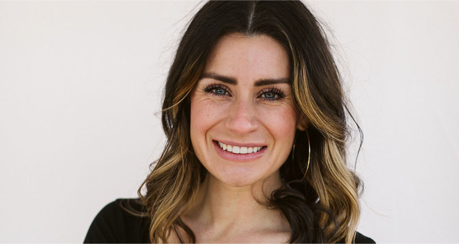 A woman with long, wavy brown hair and blonde highlights, wearing hoop earrings and a black top, smiles at the camera against a plain, light background.