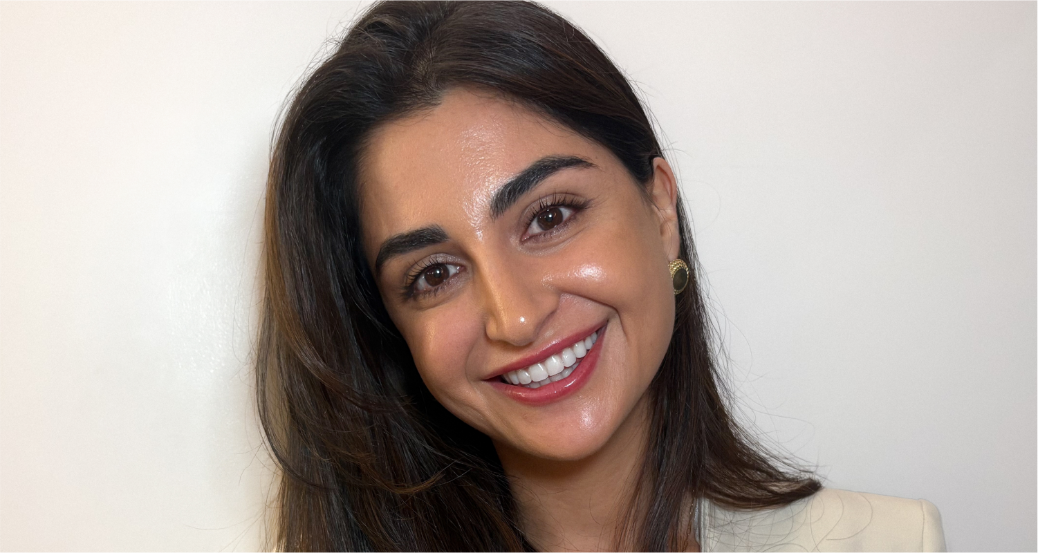 A woman with long dark hair smiles at the camera against a plain white background. She is wearing a light-colored blazer and gold earrings.