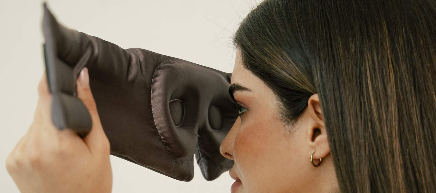 A woman with straight brown hair and gold hoop earrings holds a dark padded eye mask close to her face, preparing to put it on.