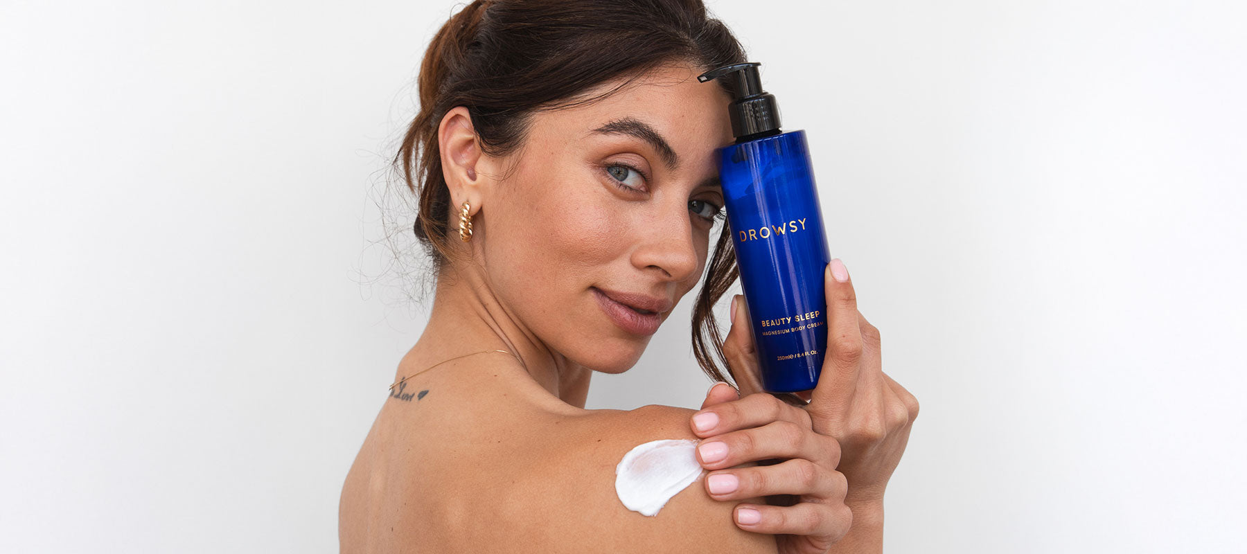 A woman with medium skin tone holds a blue bottle of Drowsy Beauty Sleep lotion, smiling slightly. She has a dollop of white lotion on her shoulder and wears gold hoop earrings. The background is plain white.