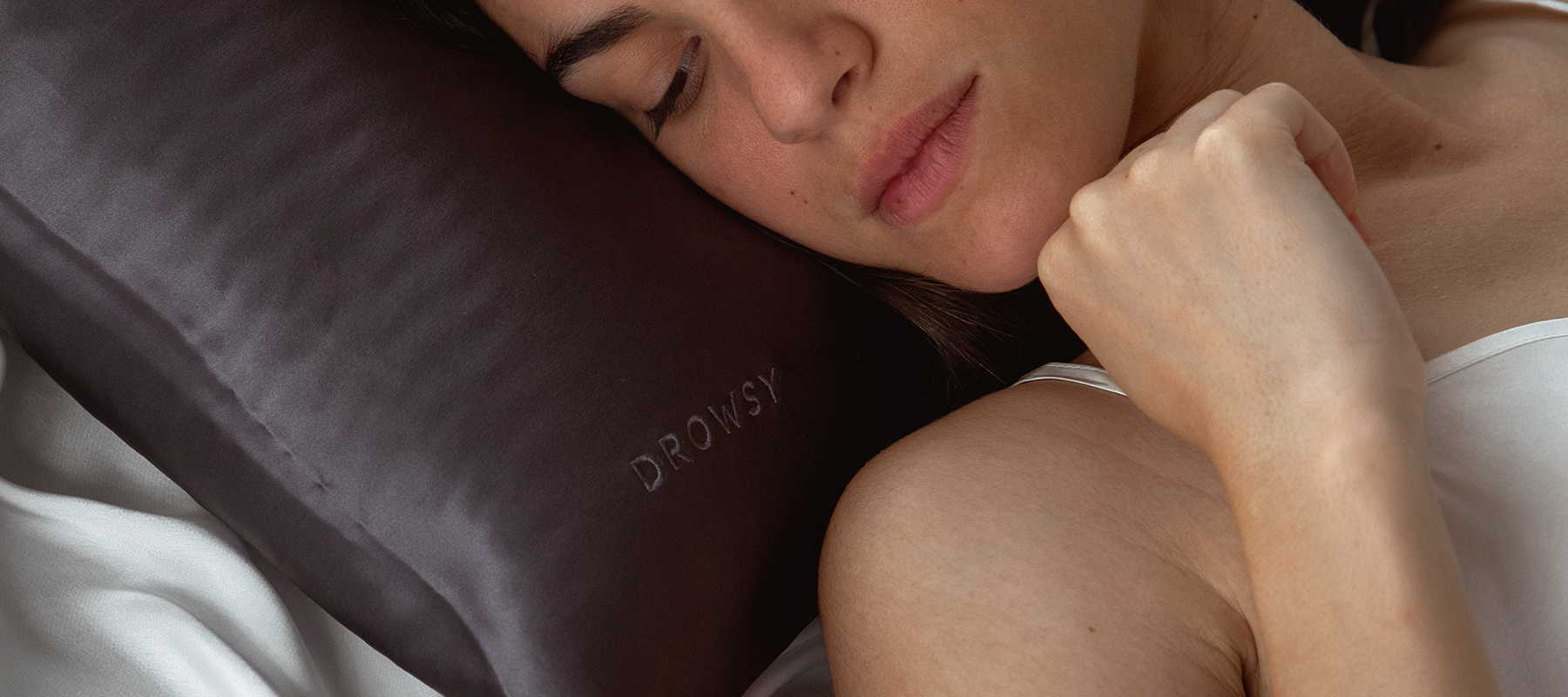 A woman in a white top sleeps on her side with her hand near her chin, resting her head on a dark gray pillow with the word DROWSY embroidered on it.