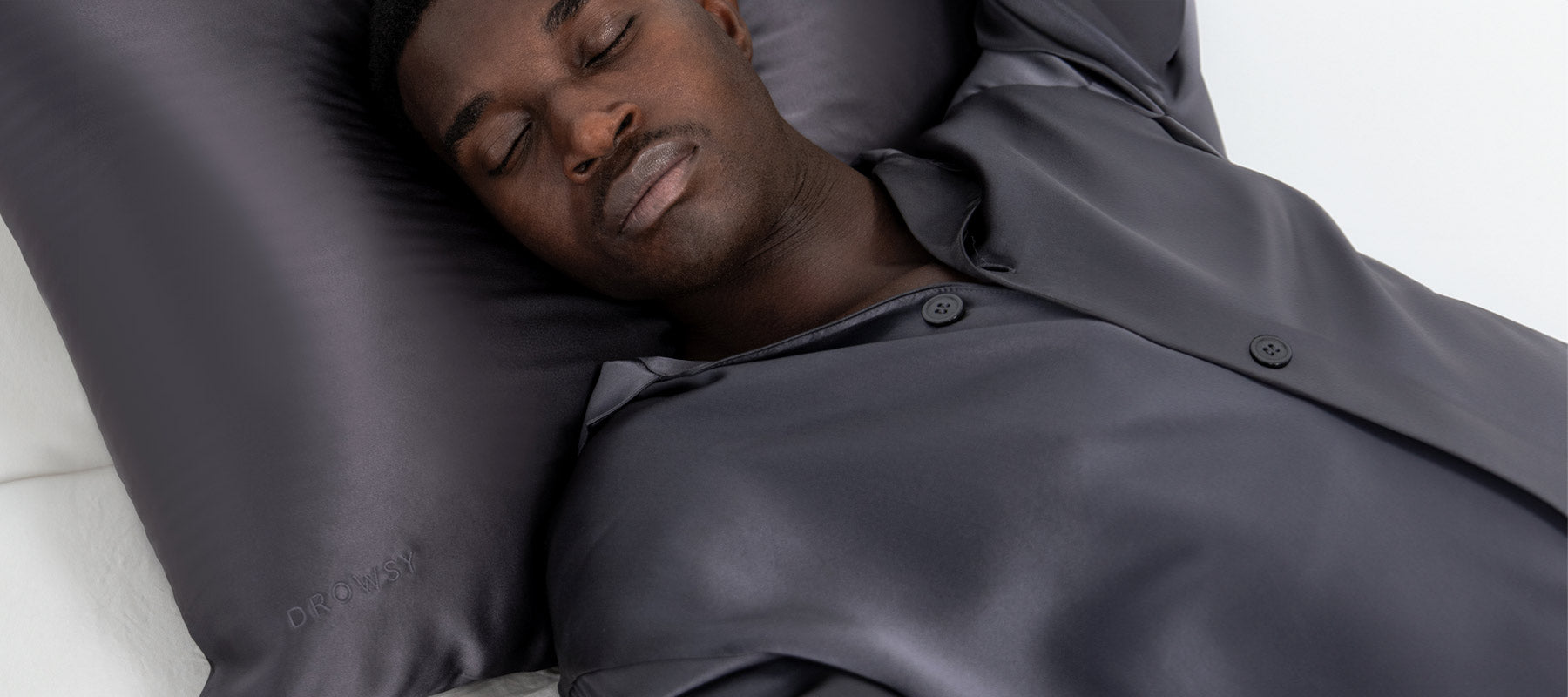 A man wearing dark satin pajamas peacefully sleeps on a matching dark satin pillow, with his arms resting above his head.