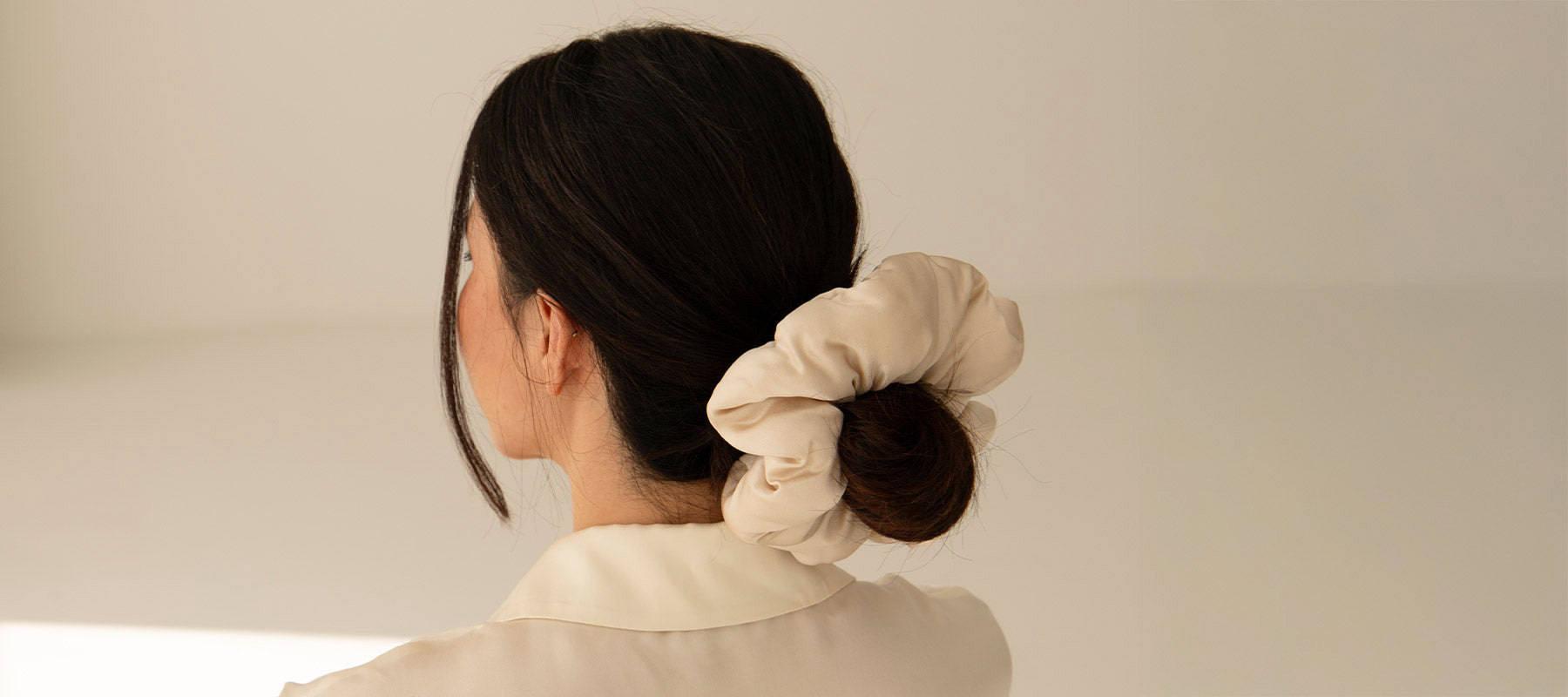 A woman with dark hair in a low bun, secured with a large beige scrunchie, is photographed from behind against a neutral background. She is wearing a light-colored, collared shirt.