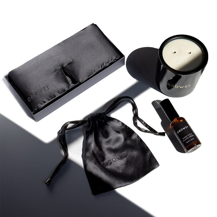 A flat lay photo features a black silk eye mask in a box, a black drawstring pouch, a black candle, and a small amber bottle with a dropper, all branded DROWSY, on a white and shadowed background.