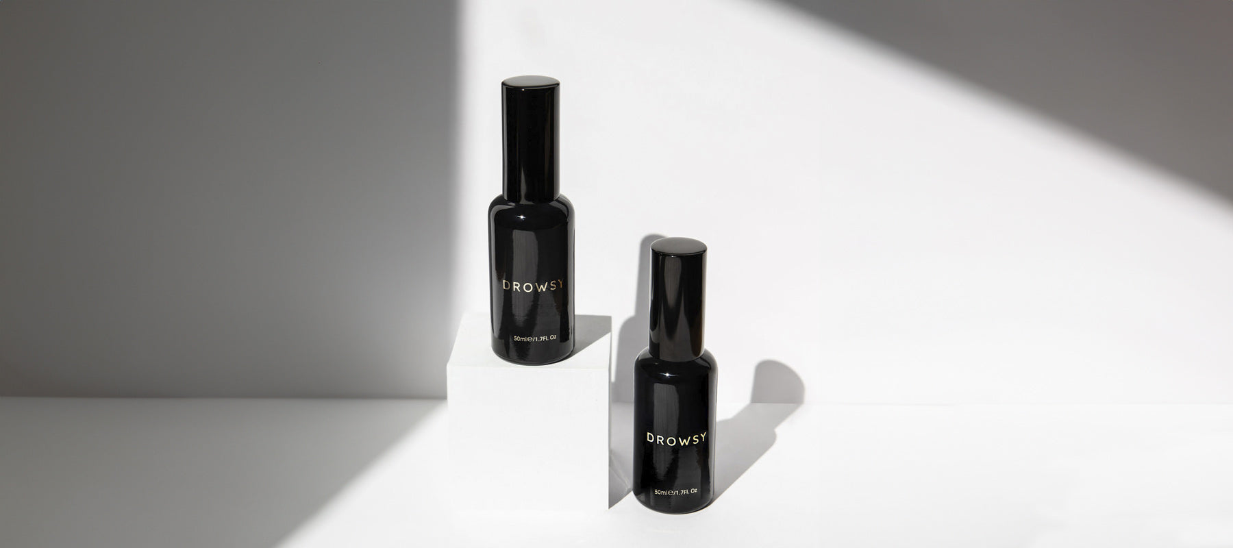 Two sleek black bottles labeled DROWSY are displayed on a white surface, with one bottle elevated on a white block. Bright light casts shadows across the minimalist background.
