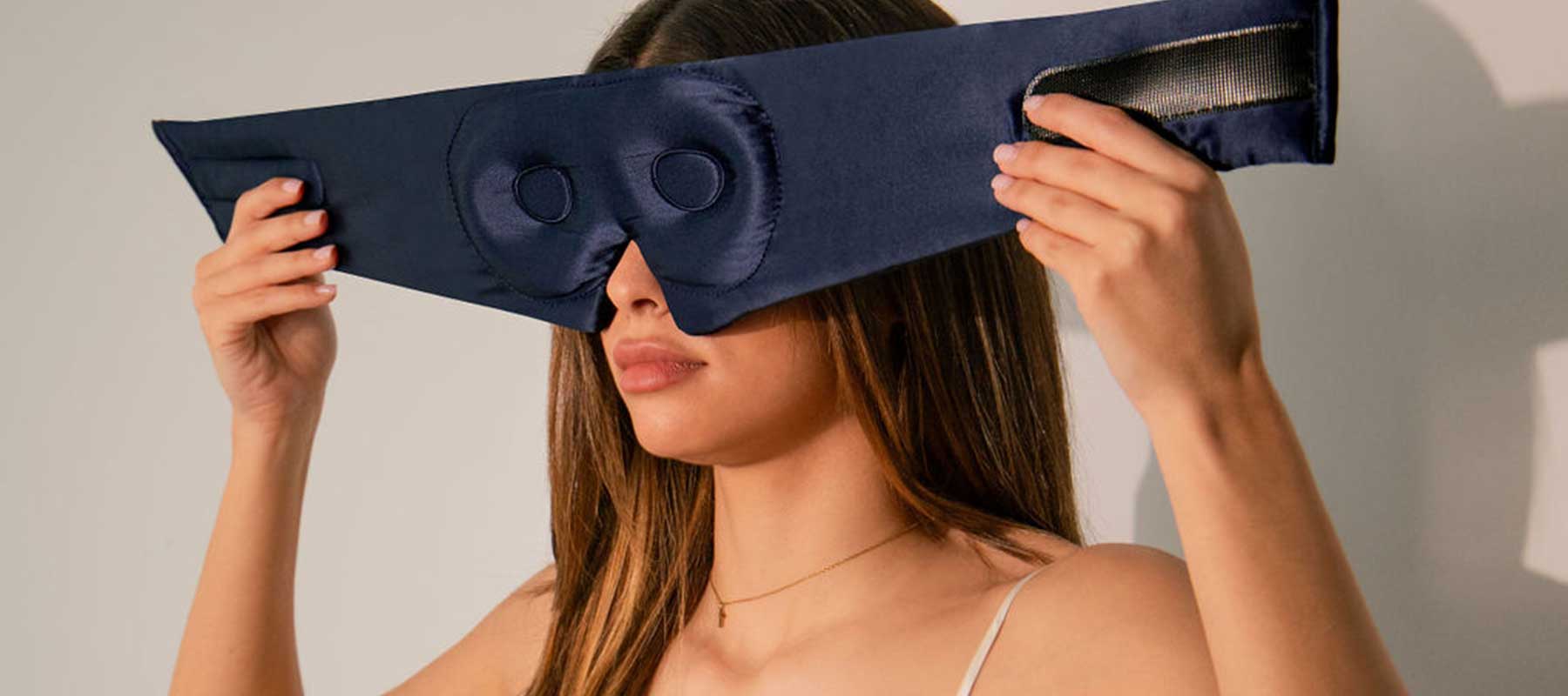 A woman with long brown hair puts on a dark blue, padded sleep mask that covers her eyes and forehead, securing it with a velcro strap around her head.