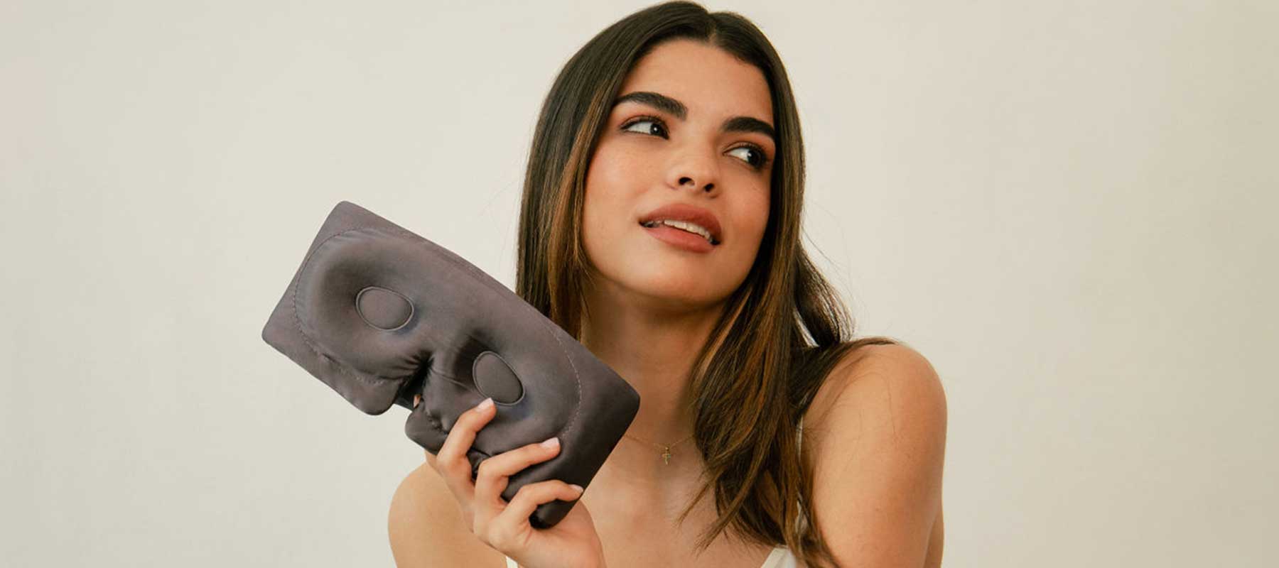 A woman with long brown hair looks to the side while holding a gray sleep mask in her hand. She wears a white top and sits against a plain, light-colored background.