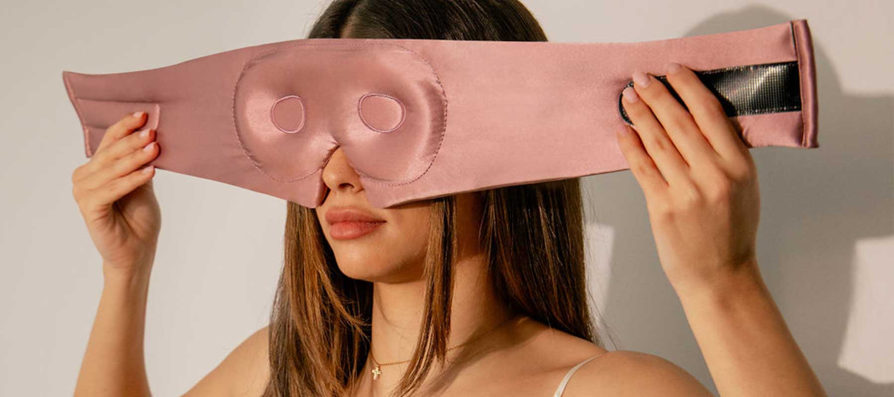 A woman holds a large, pink sleep mask with padded eye and nose areas, positioning it over her face. She has long brown hair and is wearing a white top and a delicate necklace.