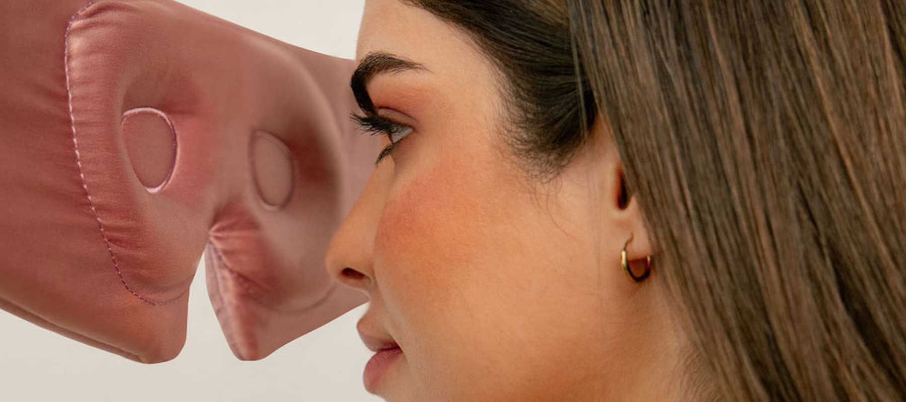 A woman with brown hair and a gold hoop earring faces a large, pink, soft fabric nose positioned close to her own nose.