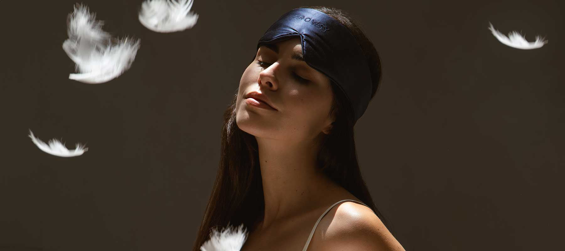 A woman wearing a black sleep mask tilts her head back with eyes closed, surrounded by floating white feathers against a dark background.