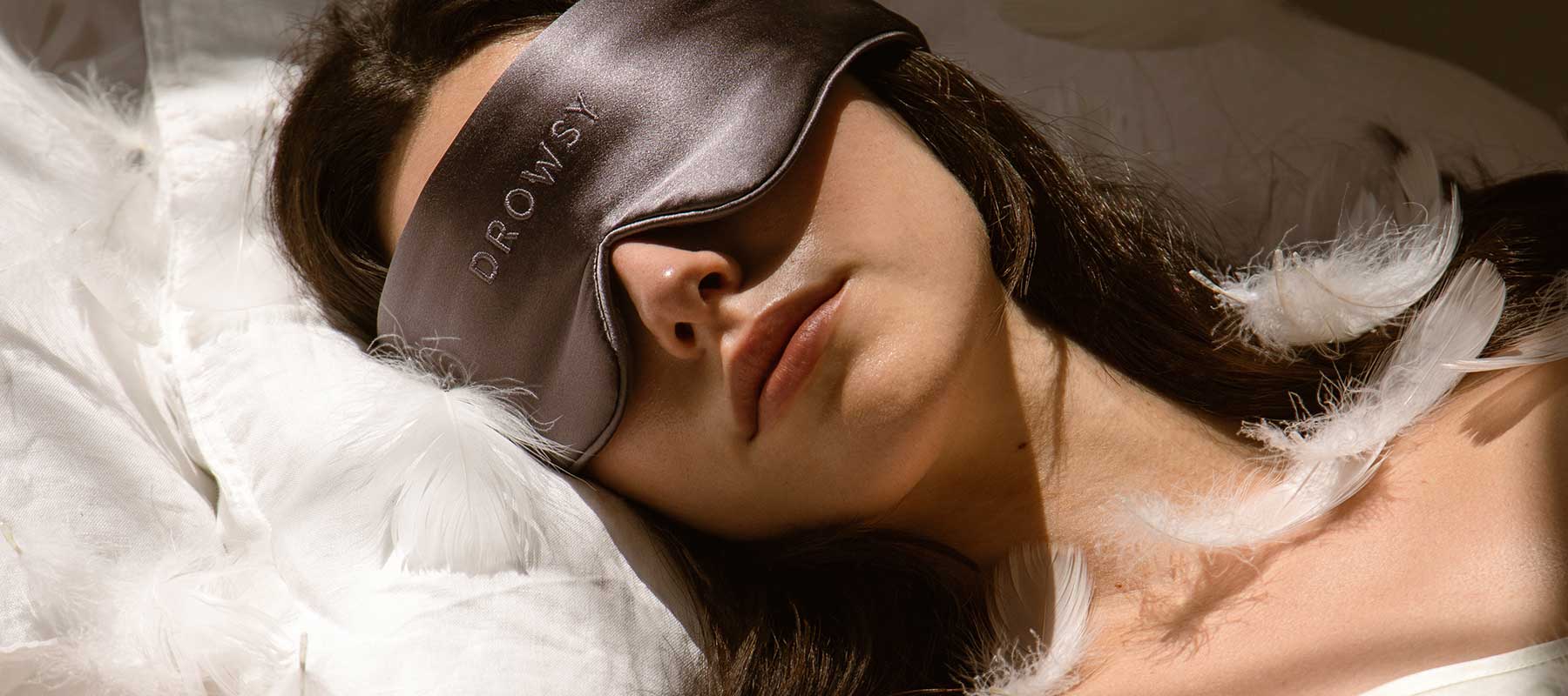 A woman lying on a white pillow with feathers, wearing a dark sleep mask that says DROWSY. She appears to be resting peacefully.