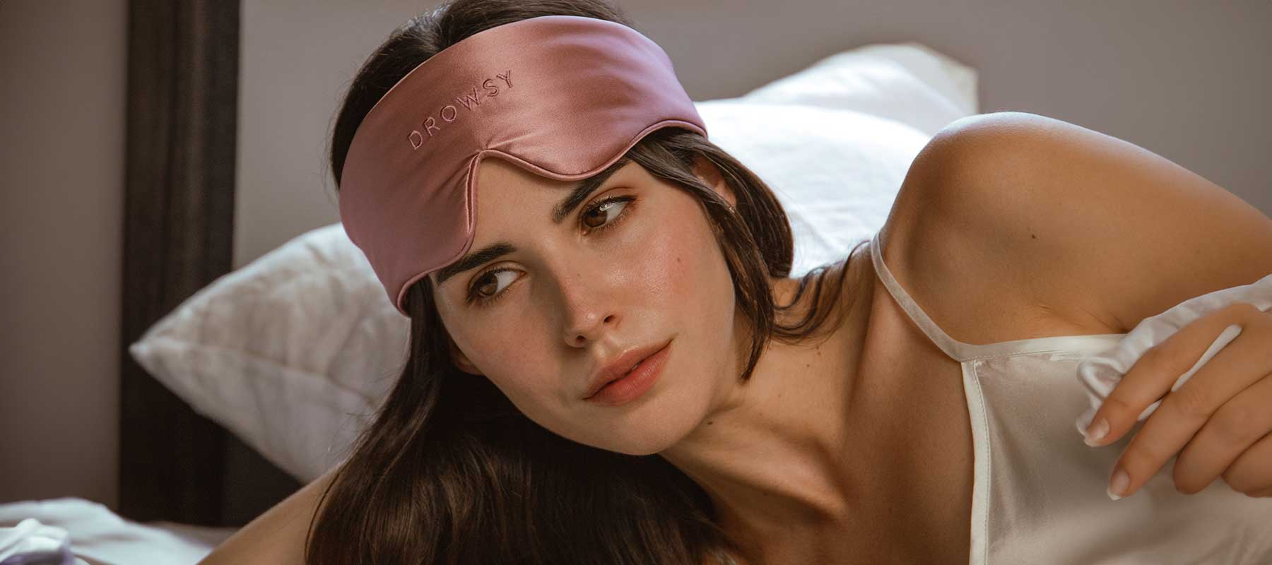 A woman in a satin camisole lies on a bed, wearing a pink sleep mask pushed up on her forehead. The mask has the word DROWSEY embroidered on it. She looks off to the side with a relaxed expression.