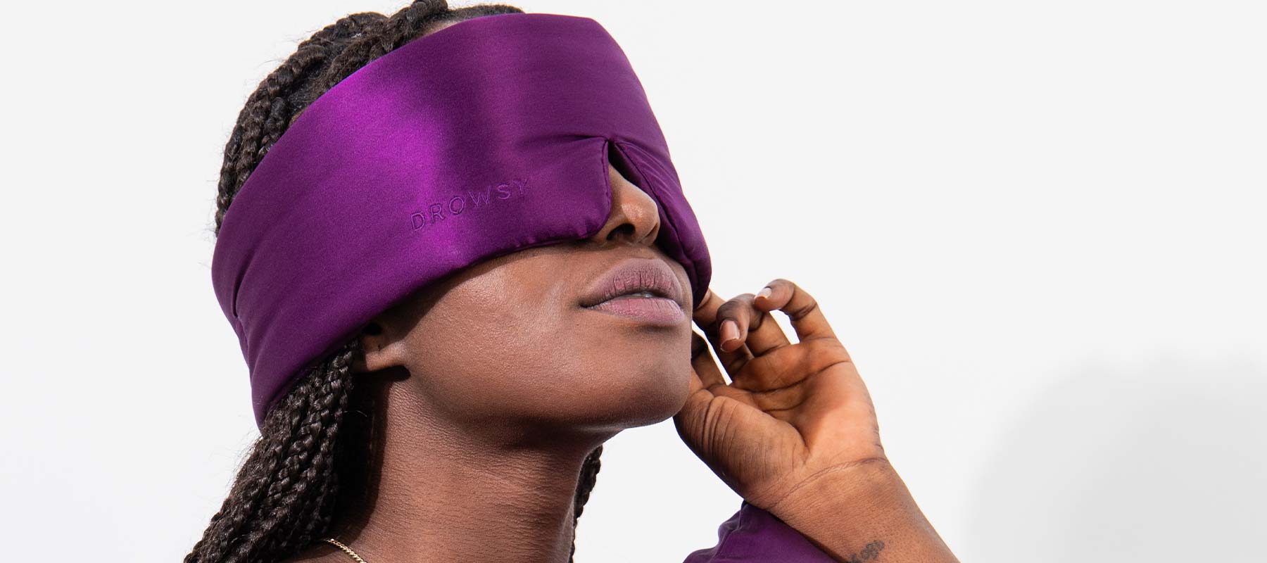 A person with braided hair wears a large, purple sleep mask covering their eyes and part of their face, holding the mask gently with one hand against a plain white background.