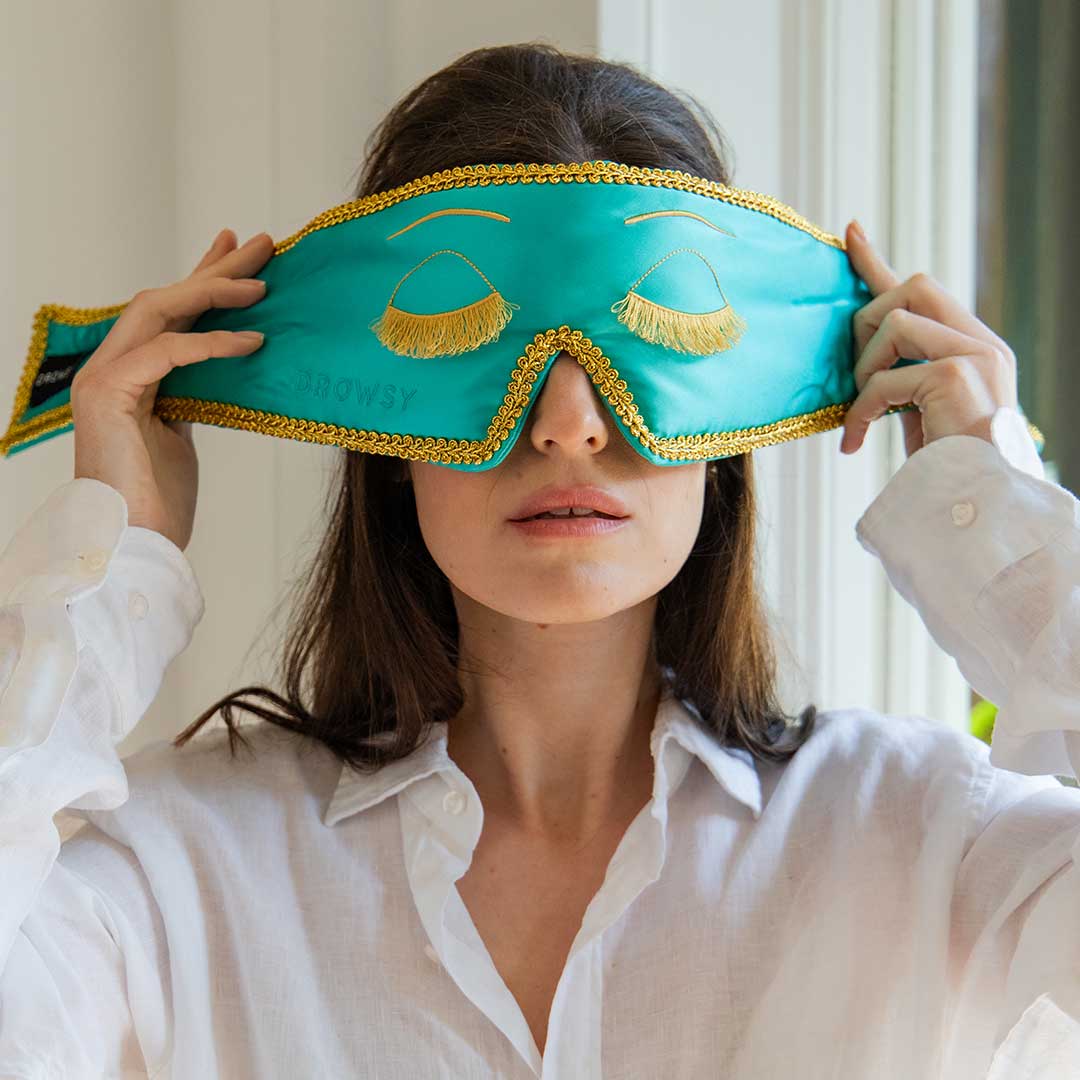 A woman wearing a white shirt holds a turquoise sleep mask with gold trim and eyelash designs over her eyes, preparing to put it on in a softly lit room.