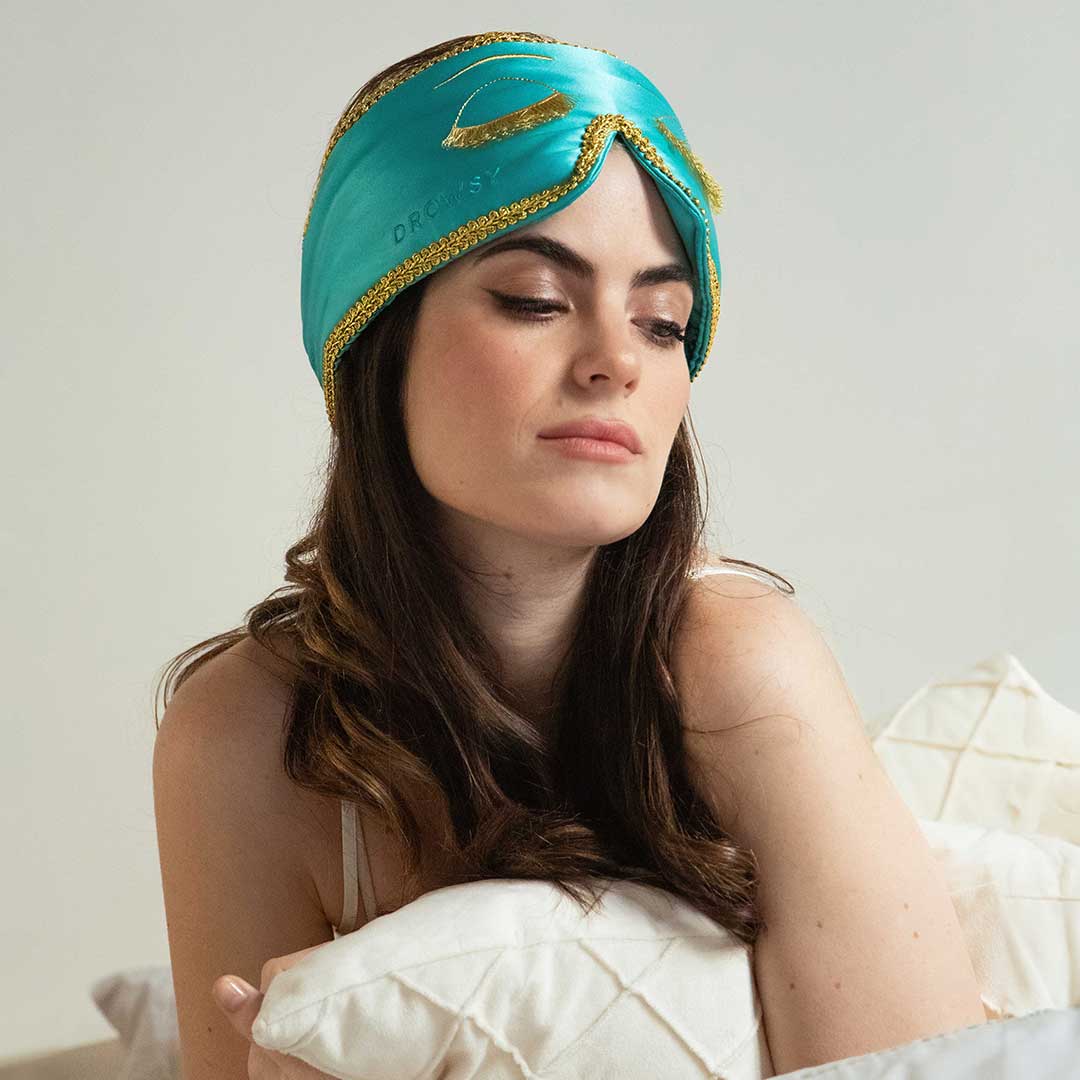 A woman with long brown hair sits on a bed, wearing a turquoise and gold sleep mask pushed up on her head. She rests her arm on a white pillow and looks down with a calm, relaxed expression.