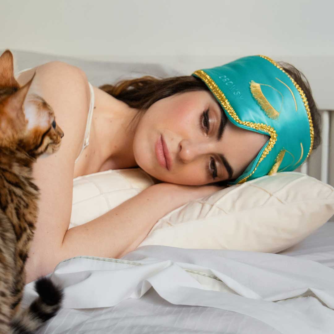 A woman wearing a green sleep mask rests on a pillow in bed, looking at a brown tabby cat sitting beside her.
