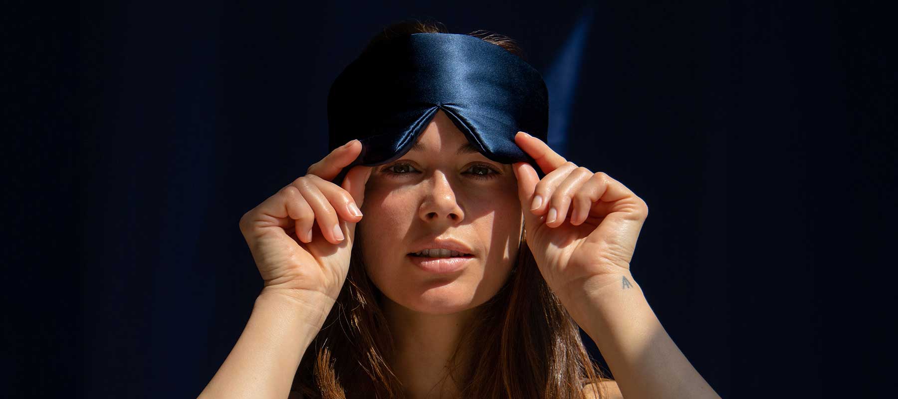 A woman with long brown hair adjusts a dark blue satin sleep mask on her forehead, standing against a dark background with sunlight illuminating her face.