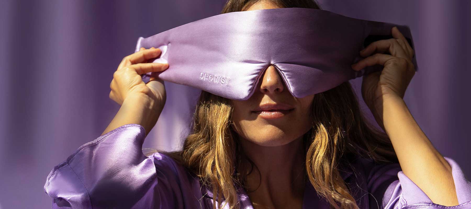 A woman wearing a shiny lavender robe adjusts a large, silky purple sleep mask over her eyes. The background is also purple, creating a calm and cohesive scene.