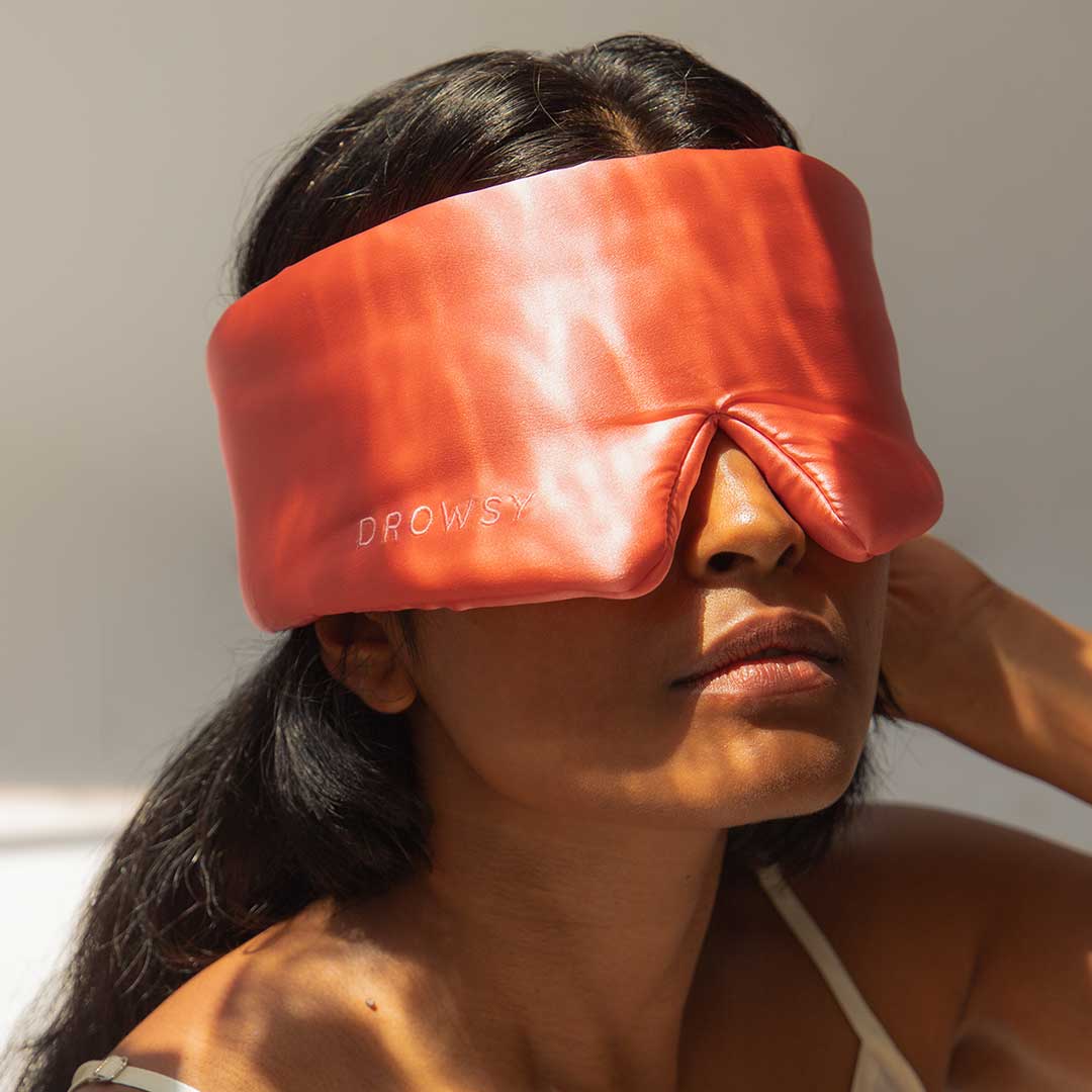 A woman wearing a wide, red silk sleep mask labeled DROWSY sits in soft sunlight, partially shadowing her face.