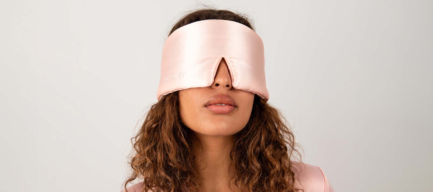 A woman with curly hair wears a light pink satin sleep mask with a front slit, covering her eyes. She stands against a plain, light-colored background.
