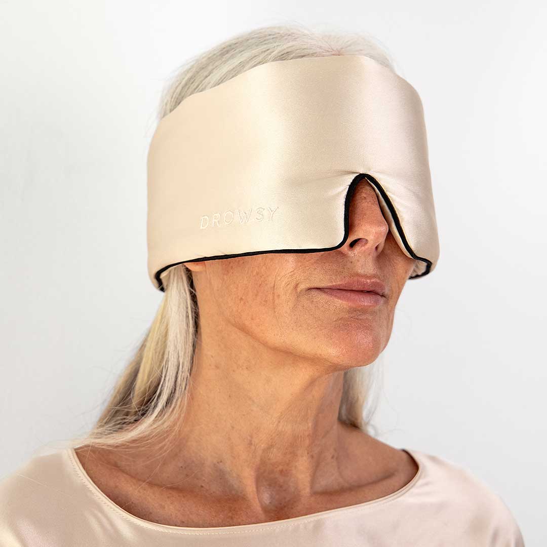 Older woman with long gray hair wearing a shiny, beige sleep mask that covers her eyes and part of her nose. The mask is labeled Drowsy and she is dressed in a matching satiny top against a plain background.