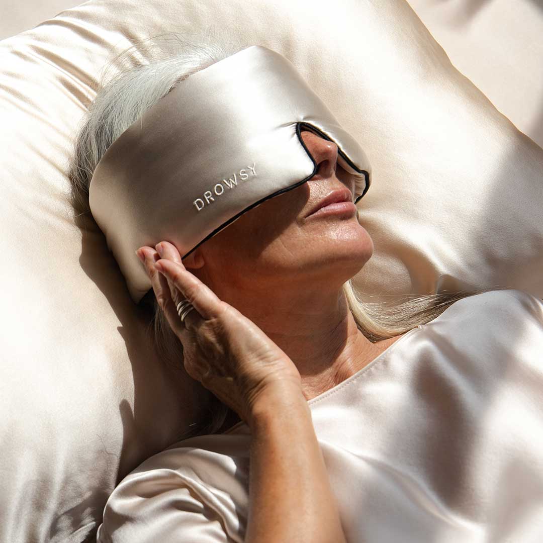 A person with long gray hair lies on a satin pillow, wearing a shiny sleep mask labeled Drowsy and a matching satin top, with one hand touching the mask and sunlight streaming over them.