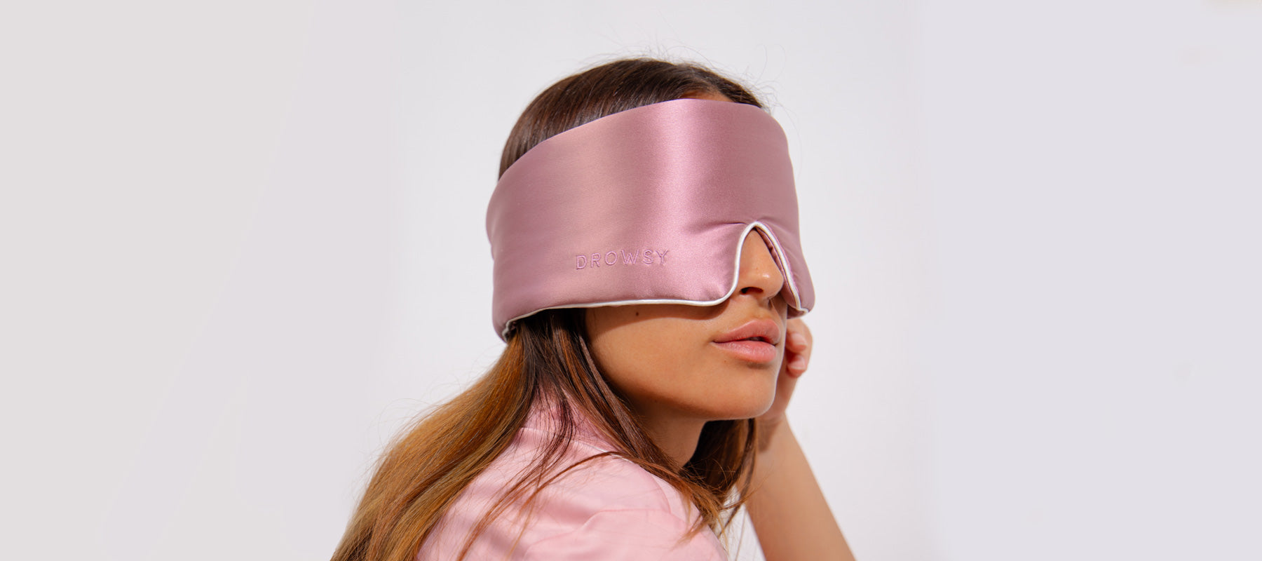 A woman with long brown hair wears a large, shiny pink sleep mask that covers her eyes and nose. She is dressed in a light pink top and faces sideways against a plain white background.