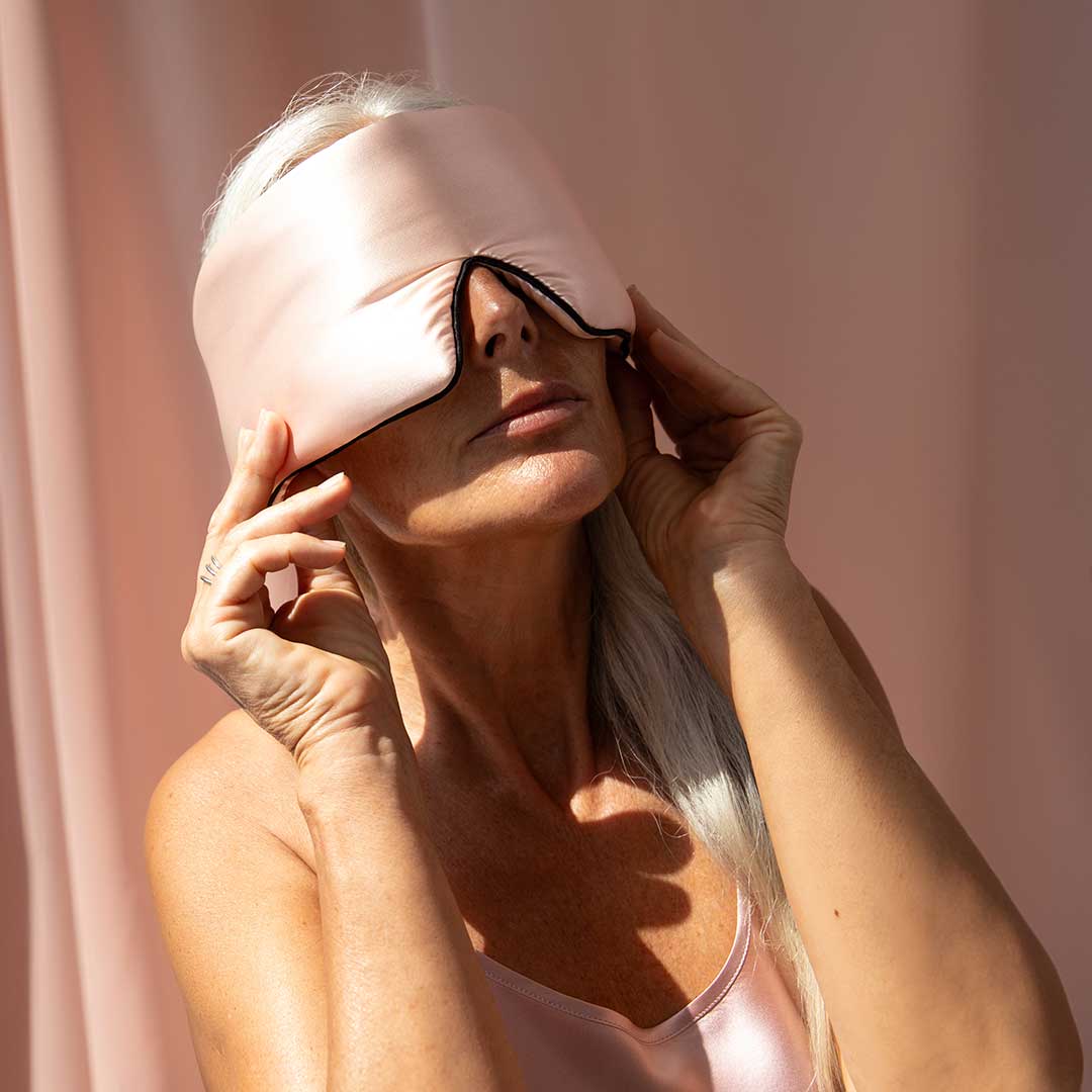 A woman with long gray hair wears a sleeveless satin top and adjusts a large pink satin sleep mask over her eyes, standing against a soft pink background.