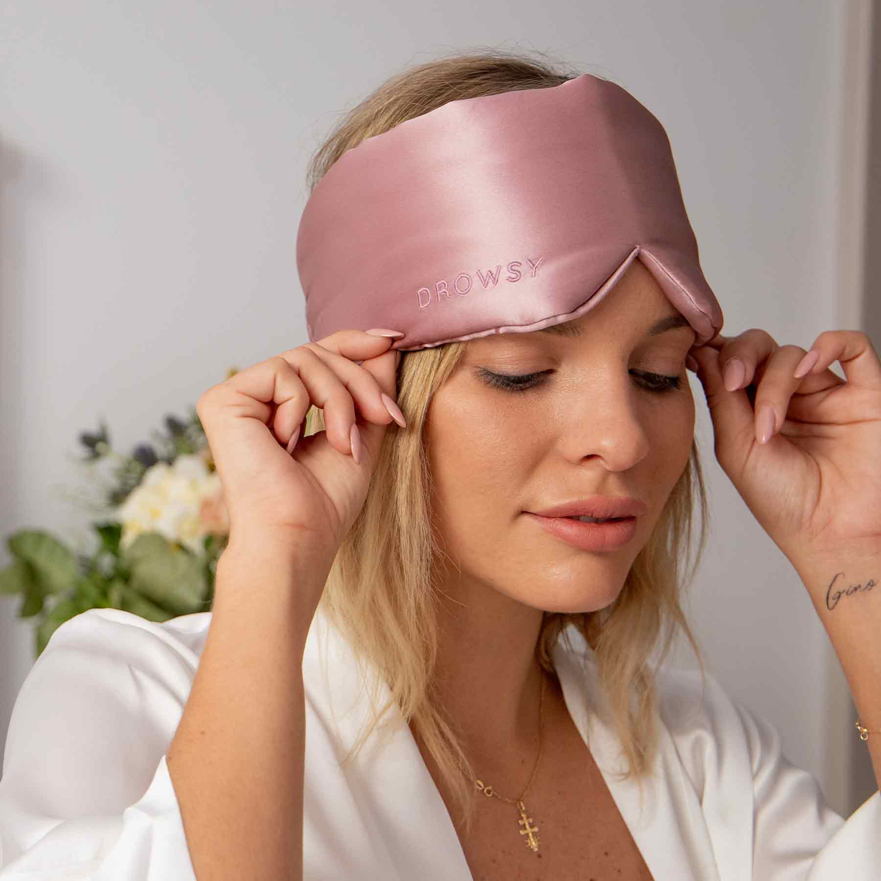 A woman with light hair in a white robe gently adjusts the Damask Rose eye mask by us.drowsysleepco on her face. A blurred bouquet of flowers is visible in the background.